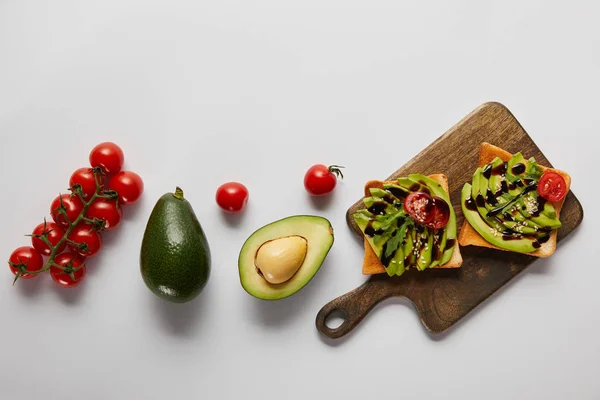 Top view of toasts on wooden cutting board with avocados and cherry tomatoes on grey backgroud — Stock Photo