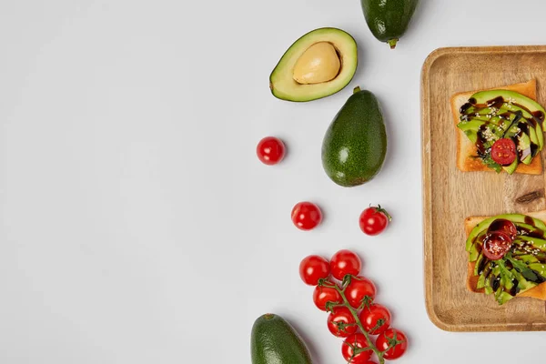 Top view of toasts on wooden cutting board with avocados and cherry tomatoes on grey backgroud — Stock Photo