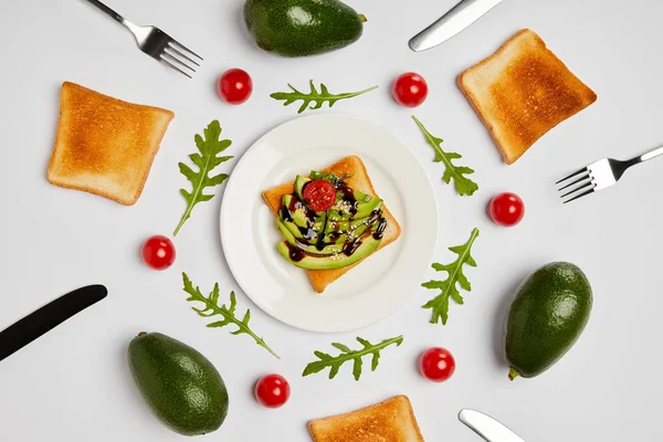 Vista superior de tostadas en plato con aguacates, tomates cherry, hojas de rúcula, tenedores y cuchillos sobre fondo gris - foto de stock