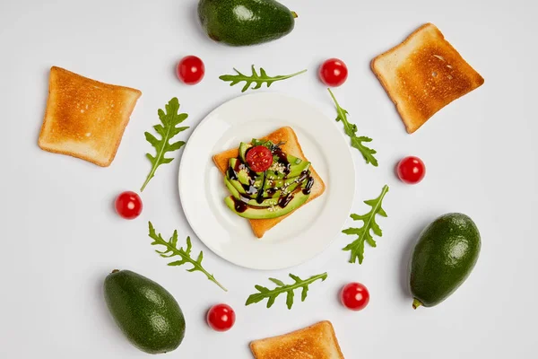 Top view of toasts on plate with avocados, cherry tomatoes and arugulas leaves on grey background — Stock Photo