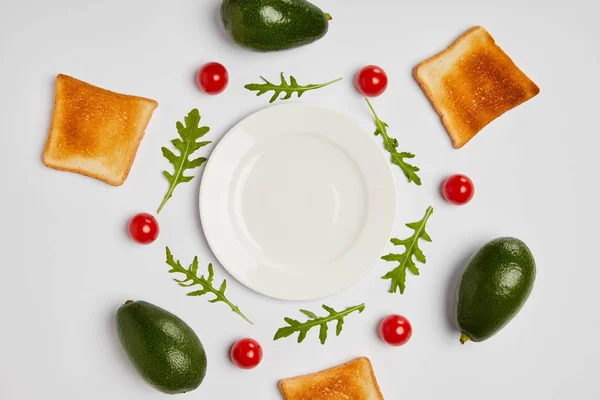 Vista dall'alto di piatti vuoti, toast, avocado, pomodorini e foglie di arugole su fondo grigio — Foto stock