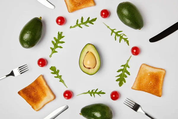 Top view of toasts, avocados, cherry tomatoes and arugulas leaves, forks and knives on grey background — Stock Photo