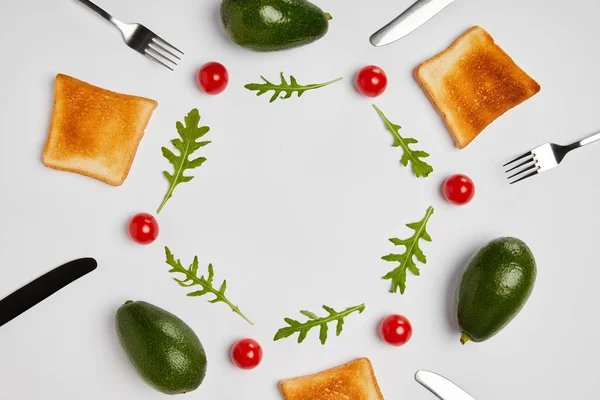 Top view of toasts, avocados, cherry tomatoes and arugulas leaves, forks and knives on grey background — Stock Photo