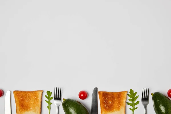 Vista dall'alto di toast, avocado, pomodorini ciliegini, foglie di arugola, forchette e coltelli su fondo grigio — Foto stock