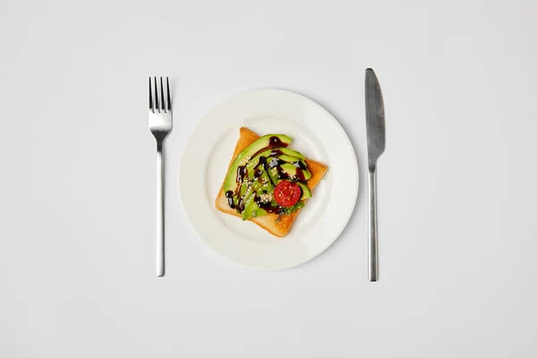 Вид зверху на тост у тарілці з авокадо та вишневим помідором, виделкою та вузлом на сірому фоні — стокове фото