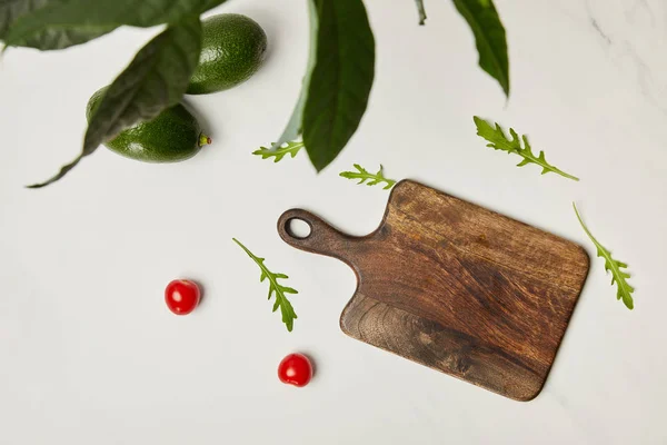 Top view of wooden cutting board, cherry tomatoes, avocados and arugula under green plant on marble surface — Stock Photo