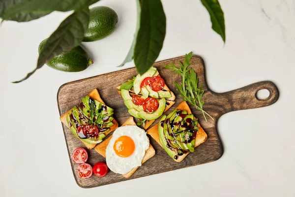 Vista superior de tabla de cortar de madera con tostadas, huevo revuelto, tomates cherry y aguacates bajo planta verde sobre superficie de mármol - foto de stock