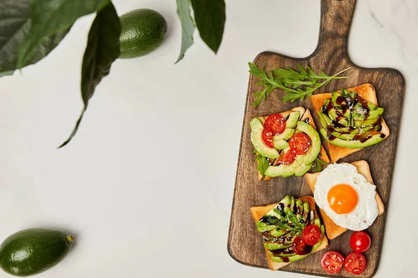 Top view of wooden cutting board with toasts, scrambled egg, cherry tomatoes and avocados under green plant on marble surface — Stock Photo