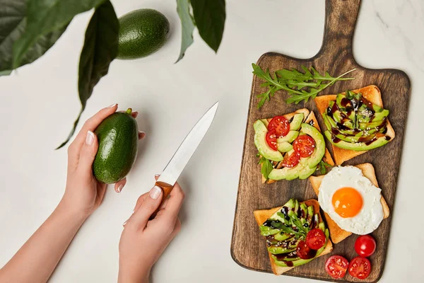 Vista dall'alto delle mani delle donne che tengono coltello con avocado e tagliere in legno con pani tostate sulla superficie di marmo — Foto stock