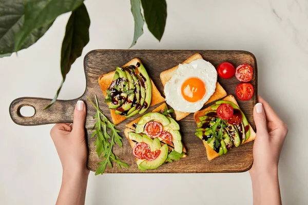 Top view of womans hands holding wooden cutting board with toasts and scrambled egg under green plant on marble surface — Stock Photo