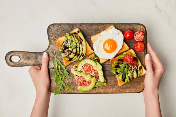 Top view of womans hands holding wooden cutting board with toasts and scrambled egg on marble surface — Stock Photo