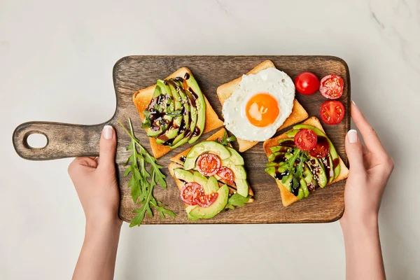 Vista superior de las manos de las mujeres sosteniendo tabla de cortar de madera con tostadas y huevo revuelto en la superficie de mármol - foto de stock