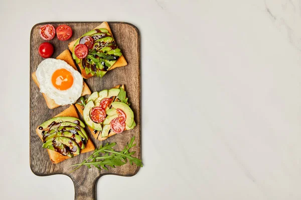 Top view of wooden cutting board with toasts, scrambled egg and cherry tomatoes on marble surface — Stock Photo