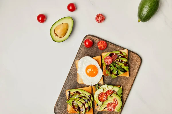 Top view of wooden cutting board with toasts, scrambled egg, avocados  and cherry tomatoes on marble surface — Stock Photo