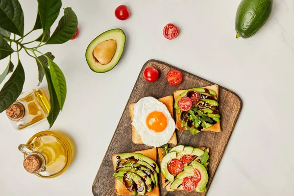 Top view of wooden cutting board with toasts, scrambled egg, cherry tomatoes, avocados and oil bottles on marble surface — Stock Photo