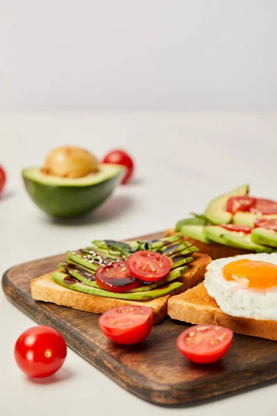 Enfoque selectivo de tabla de cortar de madera con tostadas, huevo revuelto, tomates cherry y aguacate sobre fondo gris - foto de stock