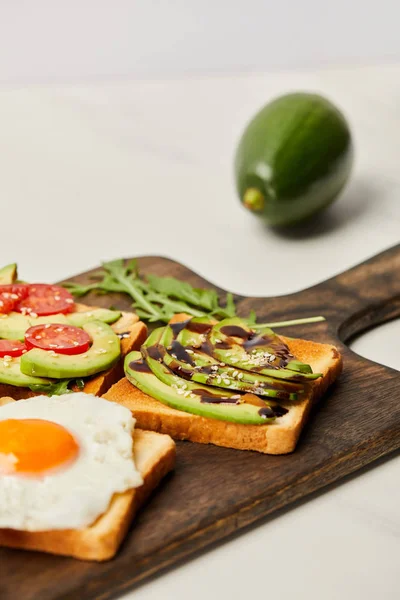 Selective focus of wooden cutting board with toasts, scrambled egg, cherry tomatoes and avocado on grey background — Stock Photo