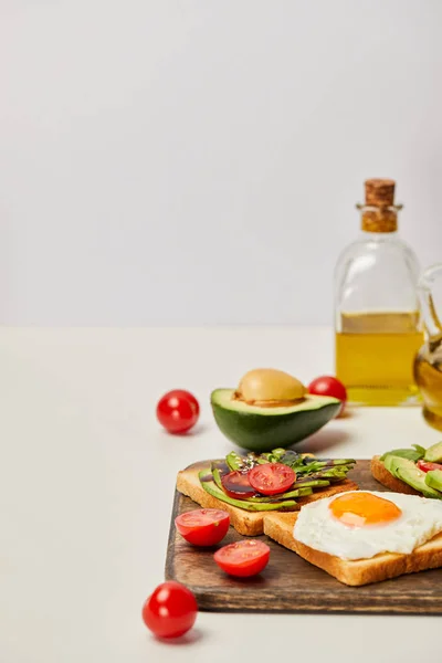 Selective focus of wooden cutting board with toasts, scrambled egg, cherry tomatoes, avocados and oil bottles on grey background — Stock Photo