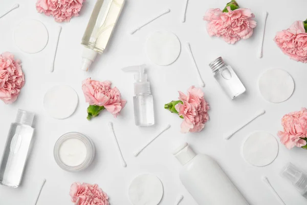 Vue du dessus de différentes bouteilles cosmétiques, fleurs d'oeillets, bâtons de coton et tampons cosmétiques sur fond blanc — Photo de stock