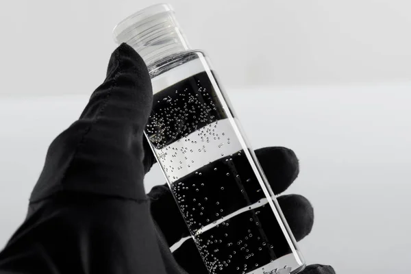 Vista cortada da mão feminina em luva preta com garrafa cosmética com líquido transparente sobre fundo branco — Fotografia de Stock