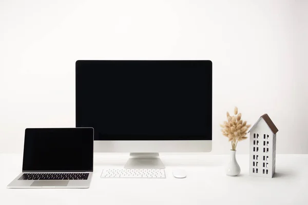 Local de trabalho com modelo de casa, flores secas em vaso, computador desktop e laptop com espaço de cópia isolado em branco — Fotografia de Stock