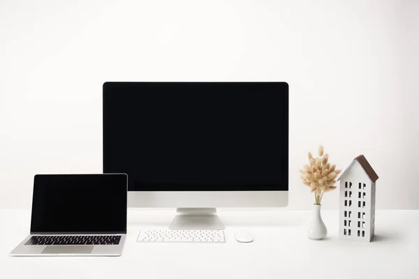 Lugar de trabajo con modelo de casa, flores secas en jarrón, computadora de escritorio y computadora portátil con espacio de copia aislado en blanco - foto de stock