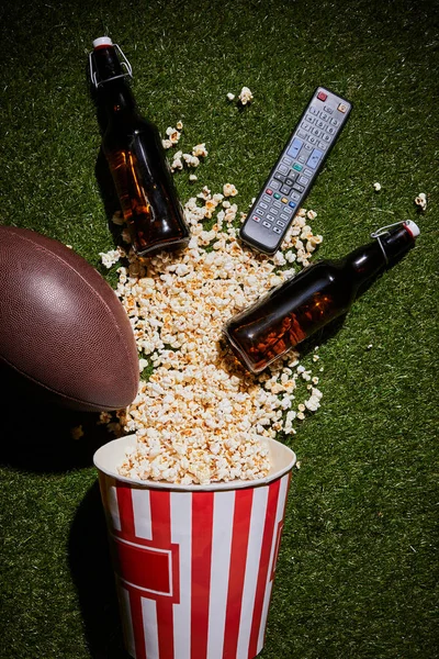 Вид коричневых бутылок рядом с попкорном, пульт дистанционного управления и мяч, лежащий на траве — стоковое фото