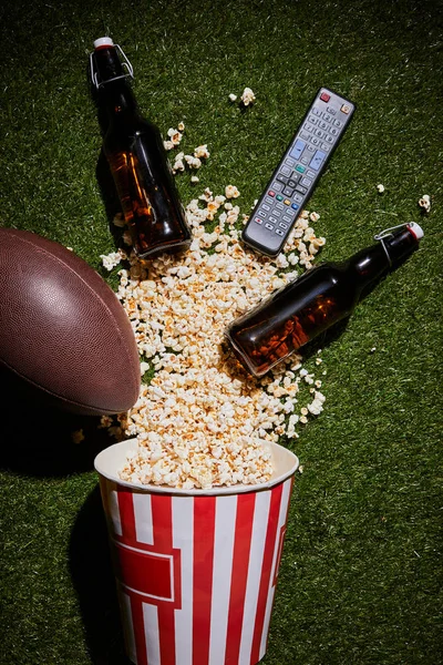 Vue du dessus des bouteilles brunes près du maïs soufflé, télécommande et balle couchée sur l'herbe — Photo de stock