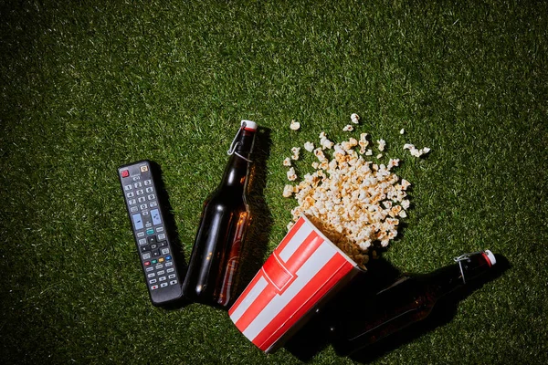Вид зверху пляшки пива біля попкорну і пульт дистанційного керування лежить на траві — стокове фото