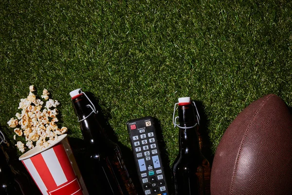 Flat lay of brown bottles near popcorn, remote control and ball lying on grass — Stock Photo