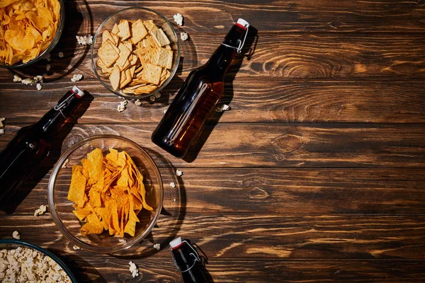 Top view of snacks in bowls near brown bottles with beer on wooden table — Stock Photo