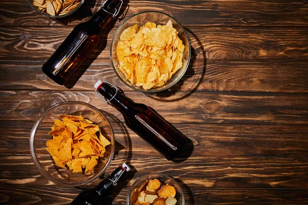 Top view of party snacks in bowls near brown bottles with beer on wooden table — Stock Photo