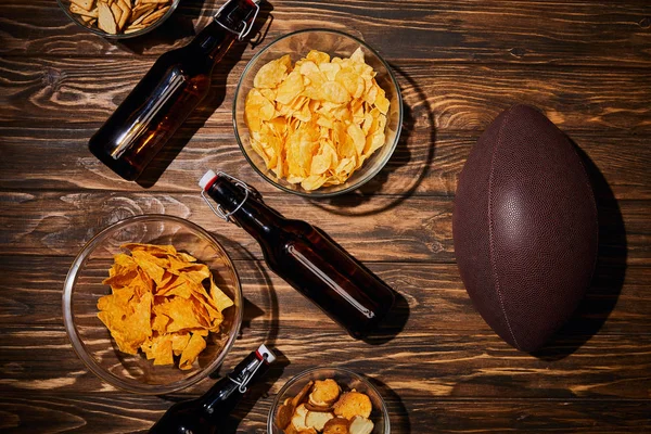 Vista superior de lanches em tigelas perto de garrafas marrons com cerveja e bola na mesa de madeira — Fotografia de Stock