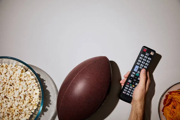 Cropped view of man holding remote control near bowls with snacks and brown ball on white background — Stock Photo