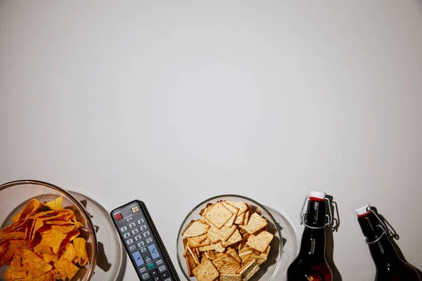Vista dall'alto di ciotole di vetro con gustosi snack vicino a bottiglie e telecomando su sfondo bianco — Foto stock