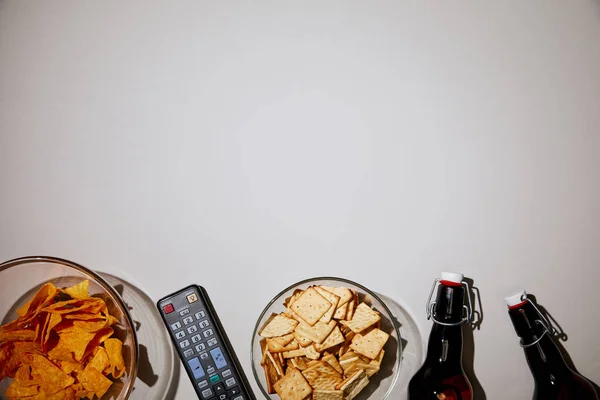 Vue de dessus des bols en verre avec des collations savoureuses près des bouteilles et télécommande sur fond blanc — Photo de stock