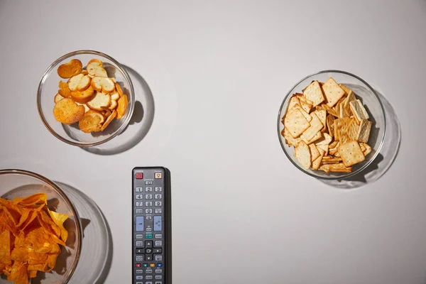 Top view of glass bowls with tasty snacks near remote control on white background — Stock Photo