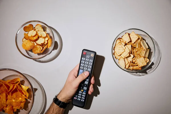 Cropped view of man holding remote control in hand near glass bowls with snacks on white background — Stock Photo