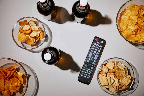Top view of bottles with beer near remote control and snacks in bowls on white background — Stock Photo