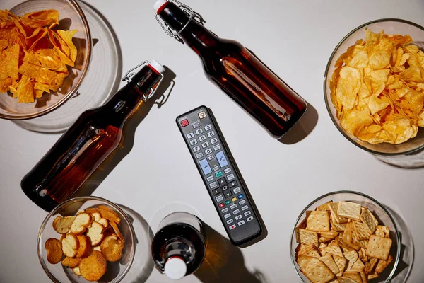 Top view of brown bottles with beer near snacks in bowls and remote control on white background — Stock Photo