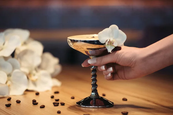 Vue recadrée d'une femme tenant un cocktail alcoolisé en verre métallique orné de fleurs d'orchidée et de grains de café — Photo de stock