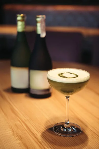 Messa a fuoco selettiva di delizioso cocktail con schiuma su tavolo di legno e bottiglie di champagne su sfondo — Foto stock