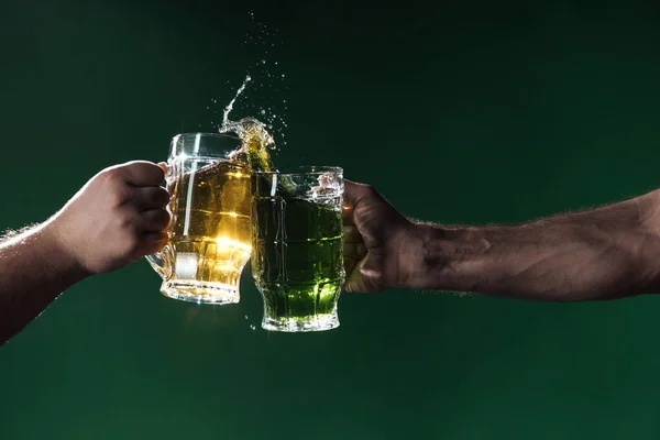 Cropped view of men clinking glasses of beer with splashes isolated on dark green, st patrick day concept — Stock Photo