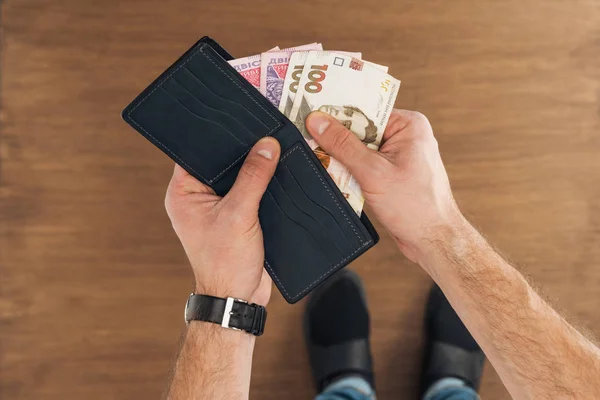 Vista superior del hombre puting billetes hryvnyas en cartera sobre fondo de madera - foto de stock