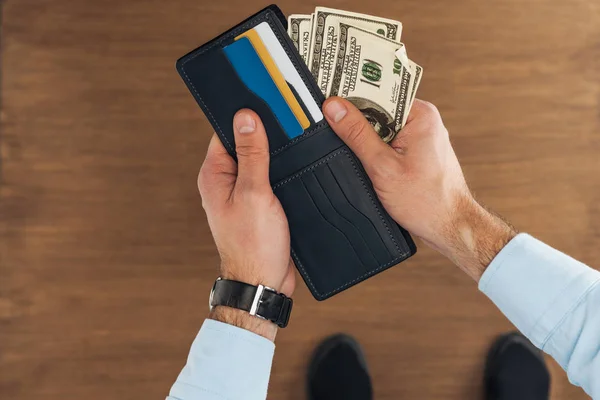 Vista superior del hombre puting billetes de dólares en cartera con tarjetas de crédito sobre fondo de madera - foto de stock