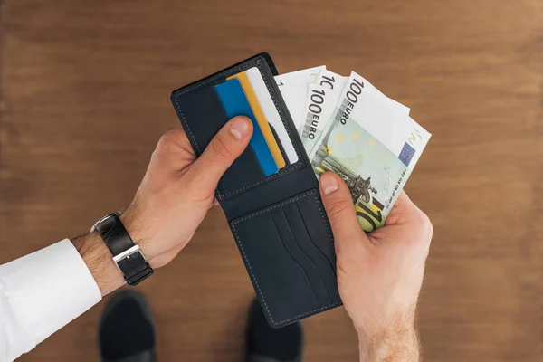 Vue du dessus de l'homme mettant des billets en euros dans un portefeuille avec des cartes de crédit sur fond en bois — Photo de stock