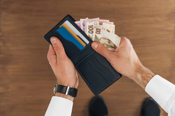 Vue du dessus de l'homme mettant des billets de banque hryvnyas dans le portefeuille avec des cartes de crédit sur fond en bois — Photo de stock