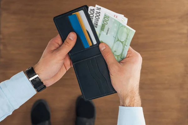 Vista superior del hombre tomando de la cartera con tarjetas de crédito billetes en euros sobre fondo de madera - foto de stock