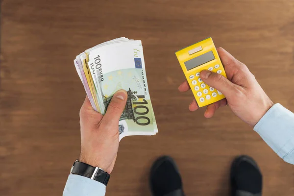 Vista superior del hombre sosteniendo billetes en euros y calculadora sobre fondo de madera - foto de stock