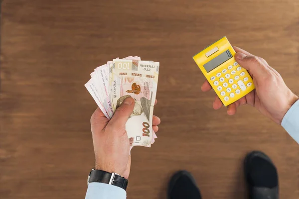 Vue de dessus de l'homme tenant des billets de banque hryvnias et calculatrice sur fond en bois — Photo de stock